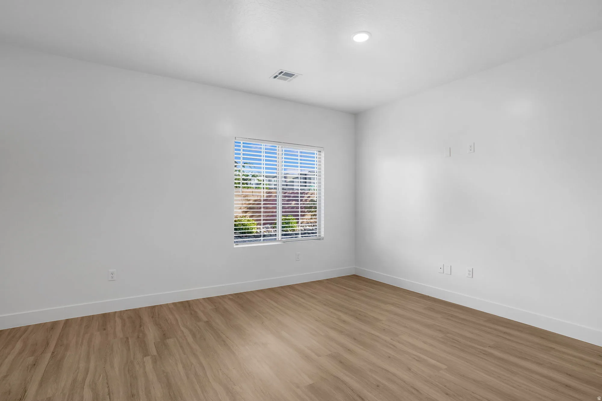 Bright and spacious bedroom showcasing pristine white walls, recessed ceiling lighting, and warm wood-finish flooring throughout
