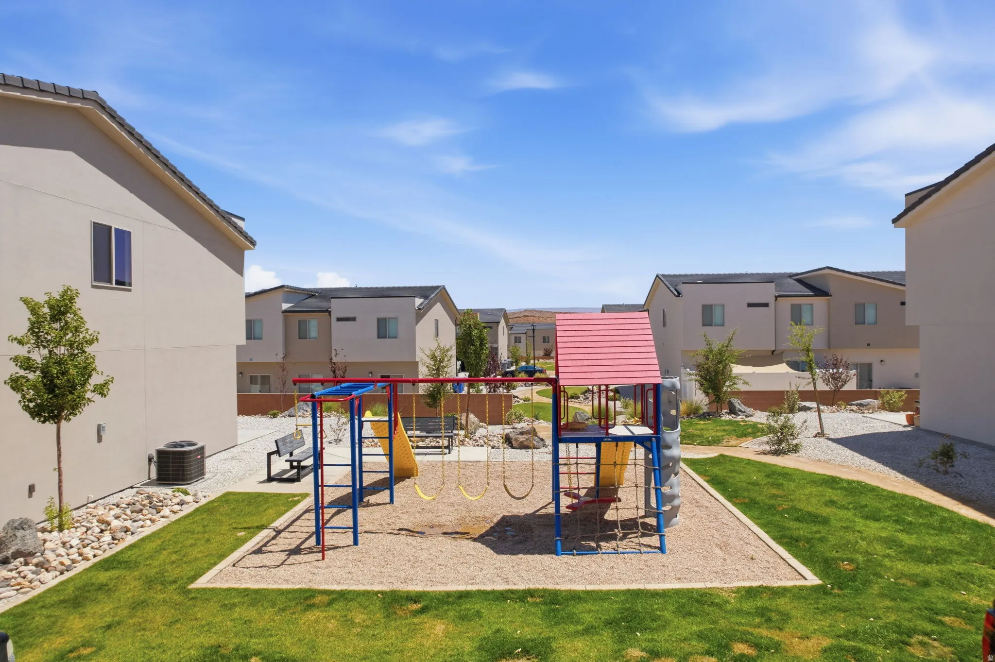 Community playground featuring a swing set, slide, and climbing structure on a wood chip surface, surrounded by green lawn and gravel landscaping