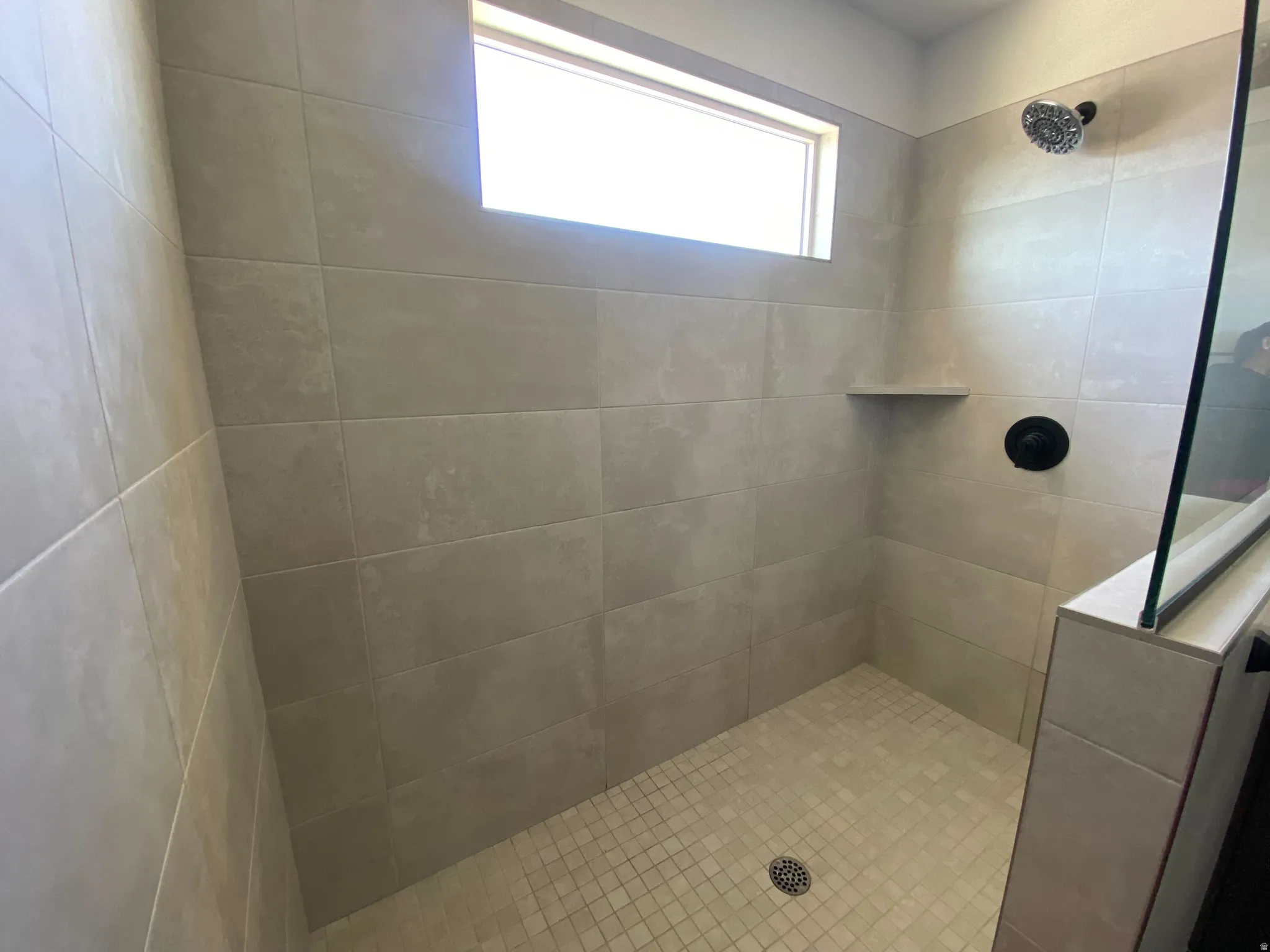 Tiled shower stall featuring a transom window, matte black fixtures, a built-in corner shelf, and a glass partition