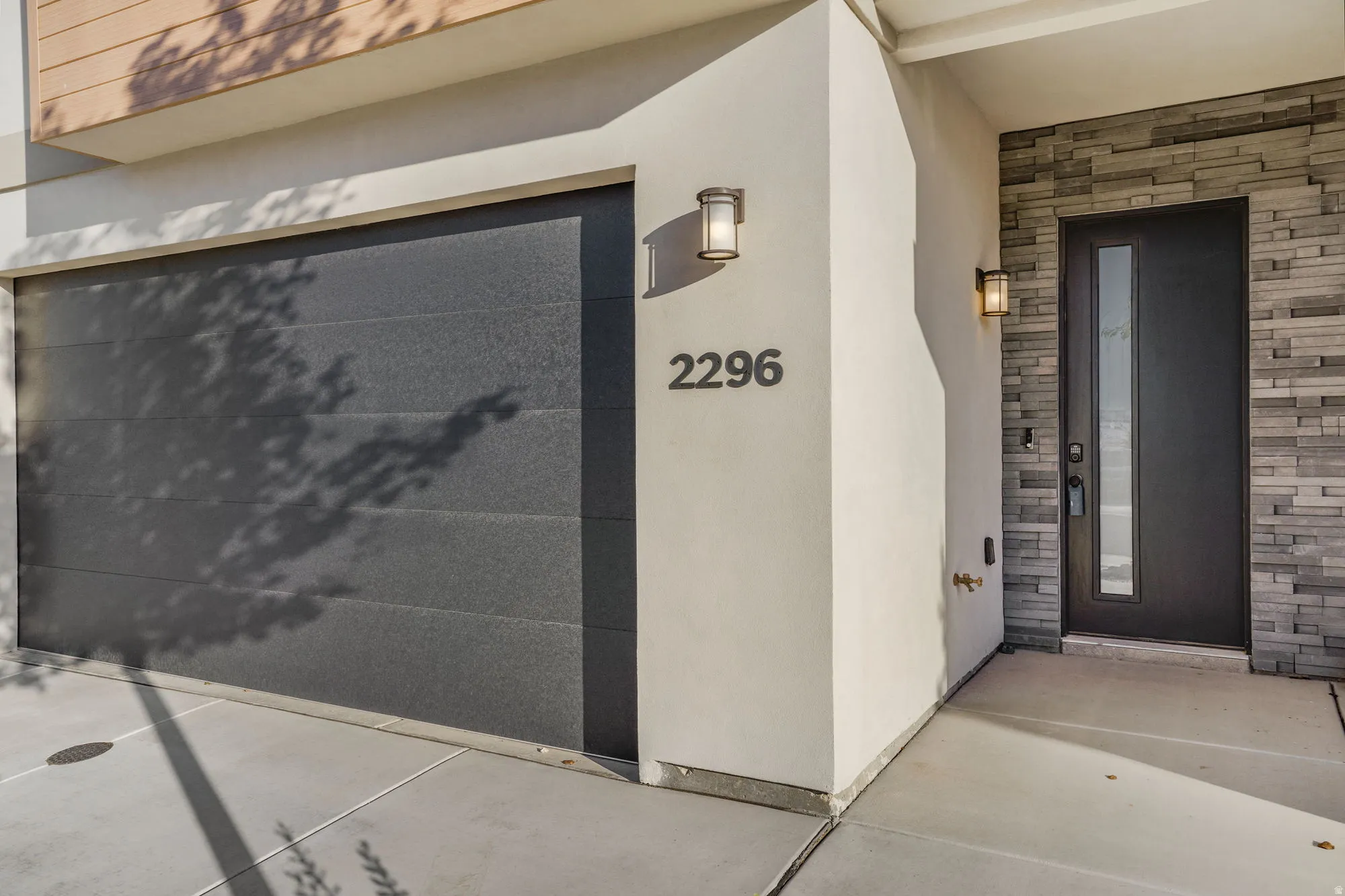 Modern exterior featuring a dark gray garage door, stacked stone accent wall, contemporary entry door with vertical glass insert, and wall-mounted sconces
