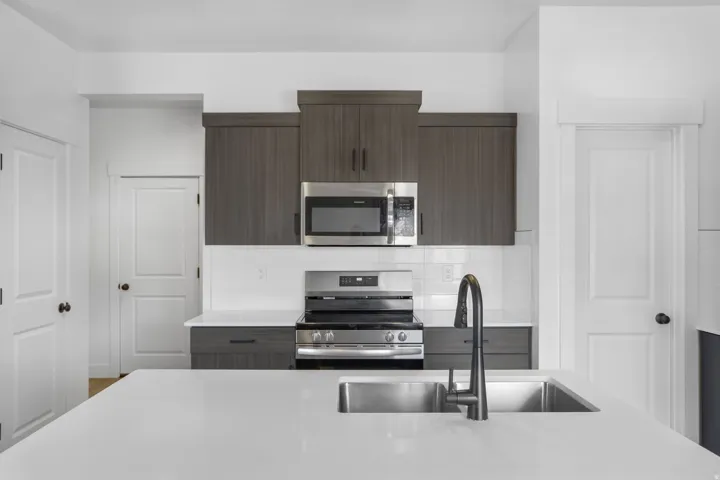 Modern kitchen featuring a white quartz countertop with waterfall edge, stainless steel undermount sink with contemporary matte black faucet, and integrated appliances including a microwave, cooktop, and wall-mounted range hood