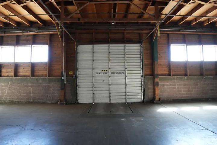 Large interior space featuring a central overhead door with a loading dock, concrete flooring, exposed wooden ceiling beams, and concrete block walls
