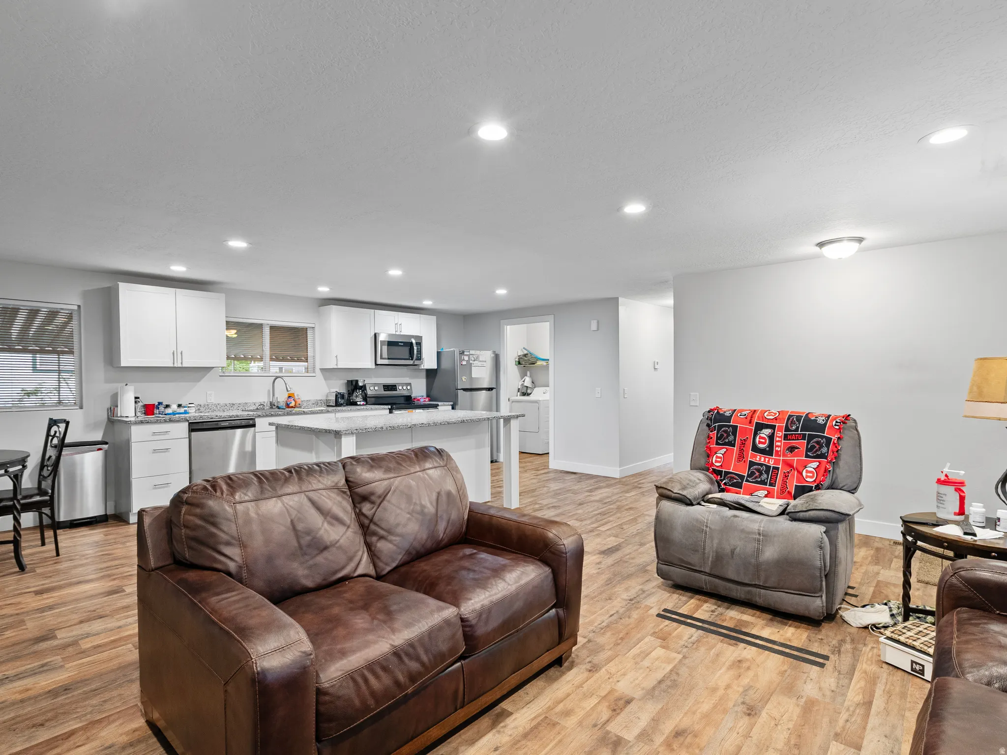 Open-concept living area featuring wood-finish flooring, recessed lighting, and a kitchen with white cabinetry, granite-style countertops, and stainless steel appliances