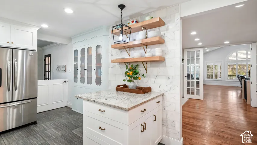 Kitchen featuring a central island with granite countertop, stainless steel refrigerator, white cabinetry, and open wood shelving on a marble-style backsplash