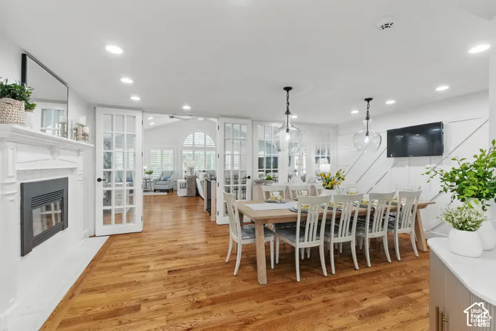 Hardwood flooring extends throughout this open concept space featuring a white mantel fireplace, french doors, a dining area with pendant lighting, and an accent wall
