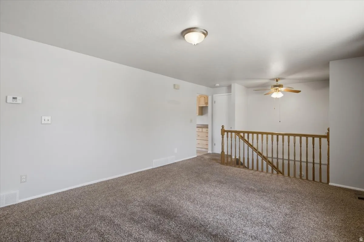 Carpeted living space featuring a ceiling fan with integrated lighting, wood railing, and a thermostat