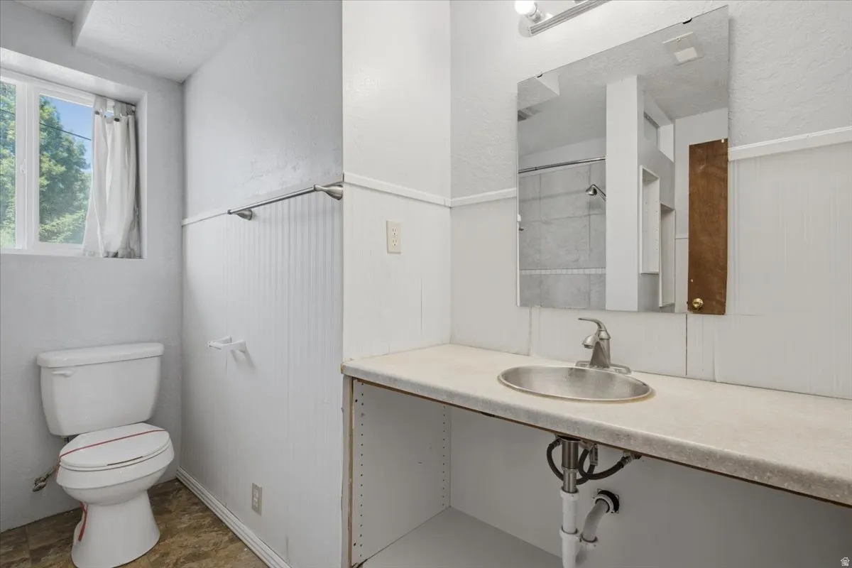 Bathroom featuring a window, white beadboard wainscoting, an oval drop-in sink, a wall-mounted mirror, and a vanity light fixture
