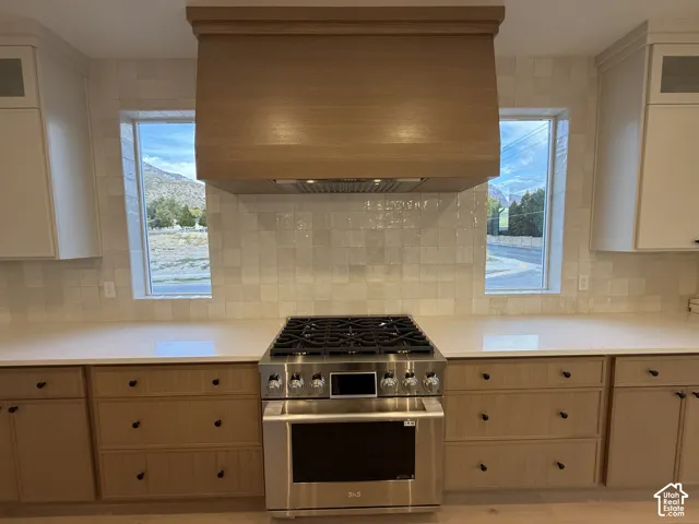 Stainless steel range, wood-finish range hood, light-toned cabinetry, light-toned countertops, and textured tile backsplash
