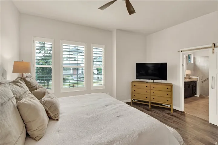 Bedroom with wood-finish flooring, a ceiling fan, and three windows featuring white plantation shutters