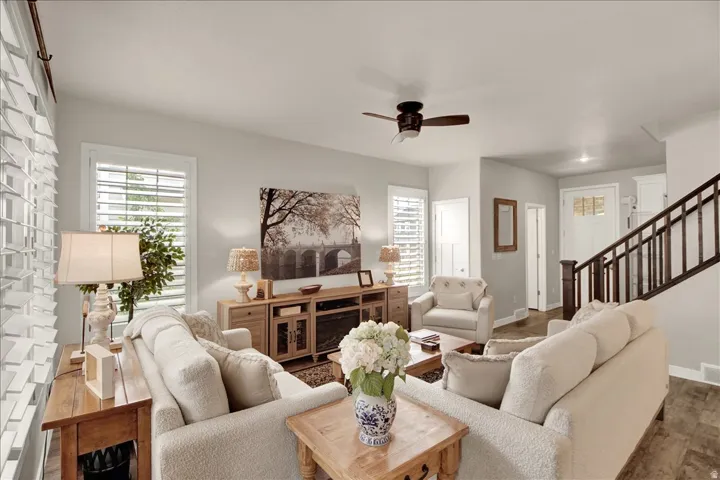 Open-concept living area featuring light-toned walls, wood-finish flooring, a ceiling fan, white window shutters, and a wood staircase with dark balusters