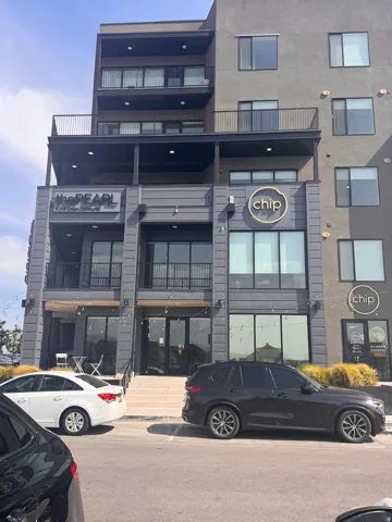 Multi-story building featuring a contemporary facade with a mix of smooth and textured finishes, multiple balconies with black railings, and prominent storefront windows on the ground level