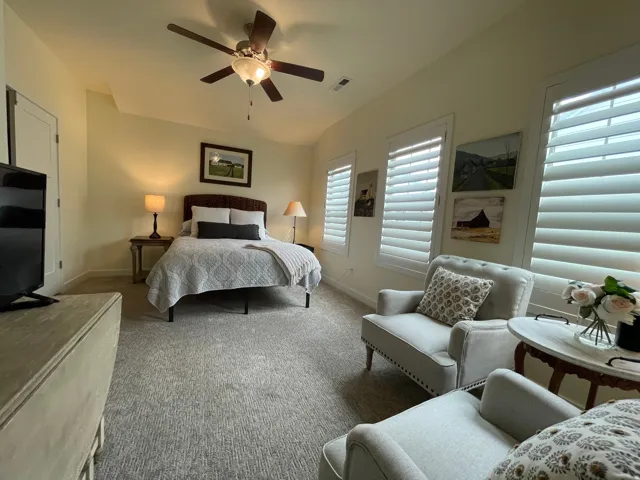 Carpeted room featuring a ceiling fan with integrated lighting, multiple windows with white plantation shutters, and a vaulted ceiling