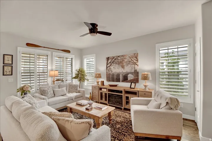 Bright living room with a ceiling fan, multiple windows featuring plantation shutters, and light wood-finish flooring