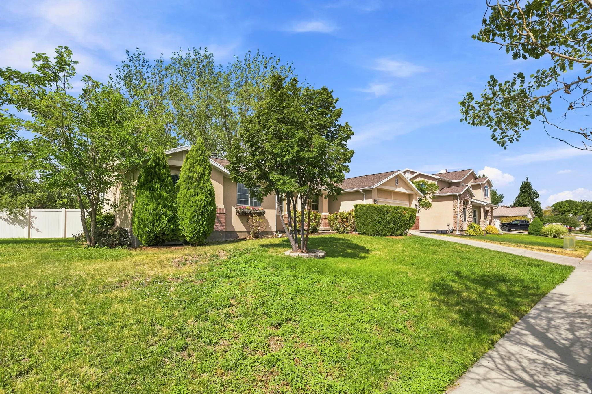 Manicured front yard featuring mature landscaping, a sidewalk, and a white privacy fence
