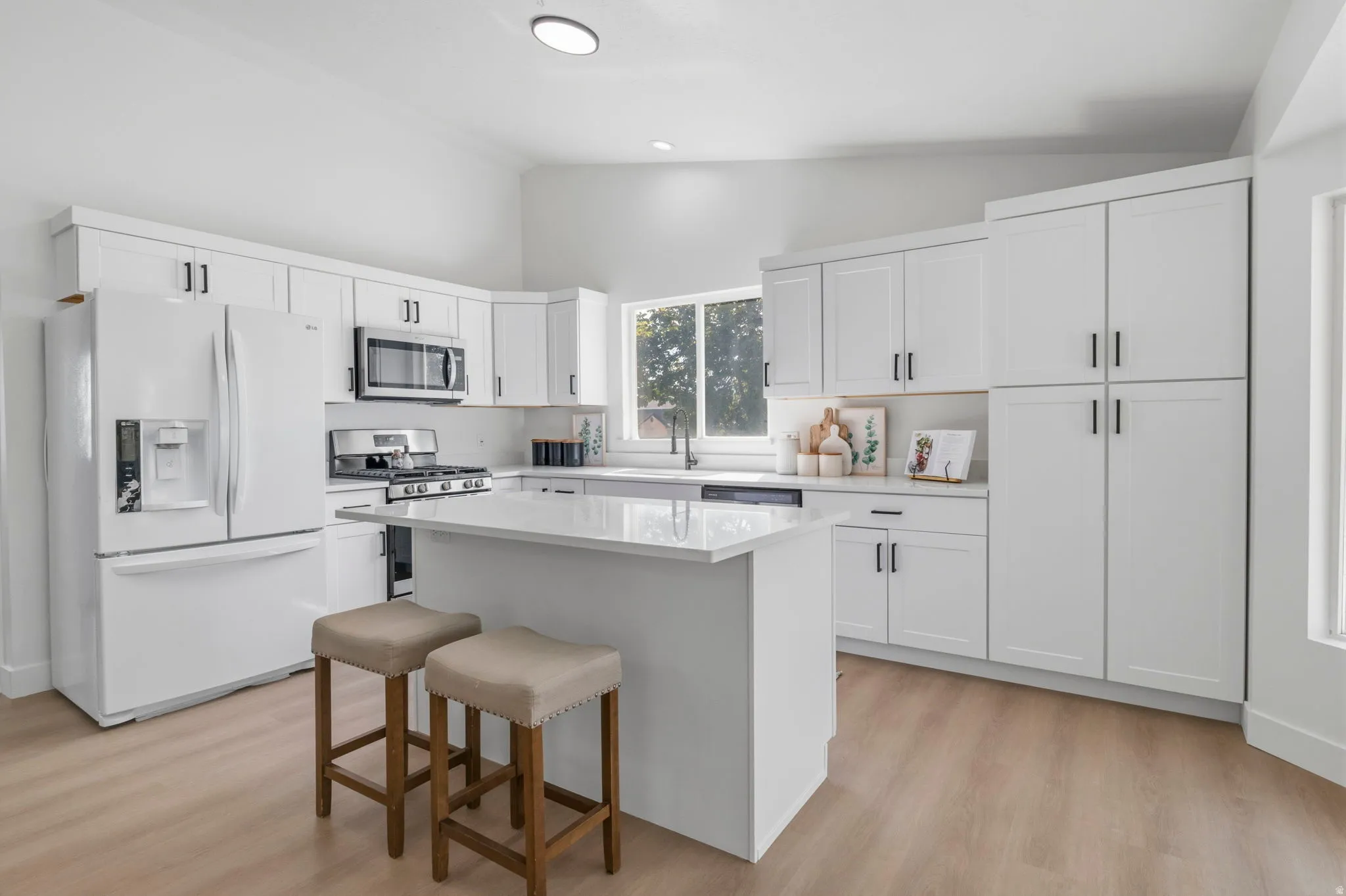 Modern kitchen featuring white shaker cabinetry, stainless steel appliances, a center island with a countertop, wood-finish flooring, and a large window