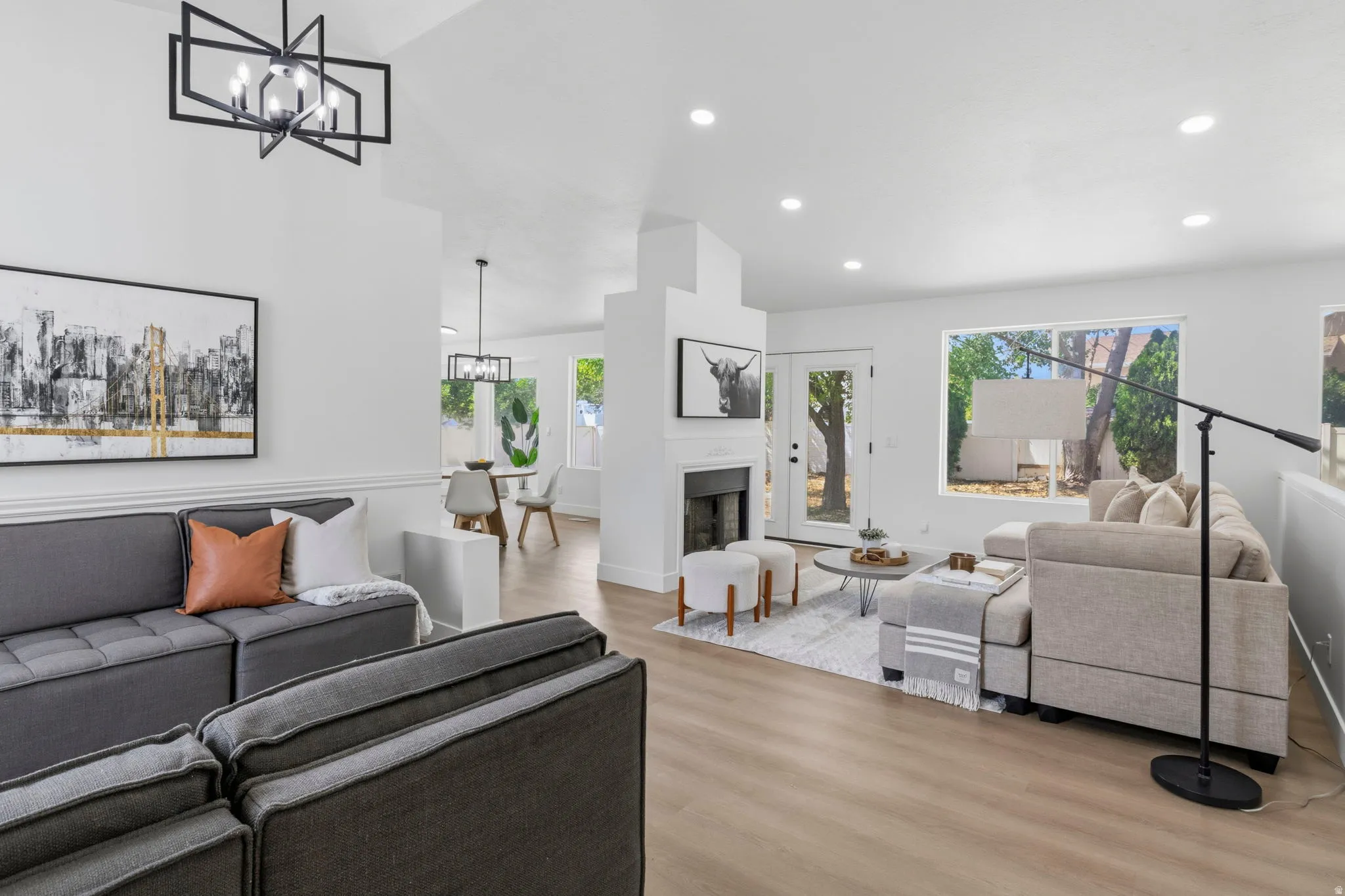 Spacious living area featuring wood-finish flooring, a contemporary black geometric chandelier, recessed lighting, and a white brick fireplace with a dark insert