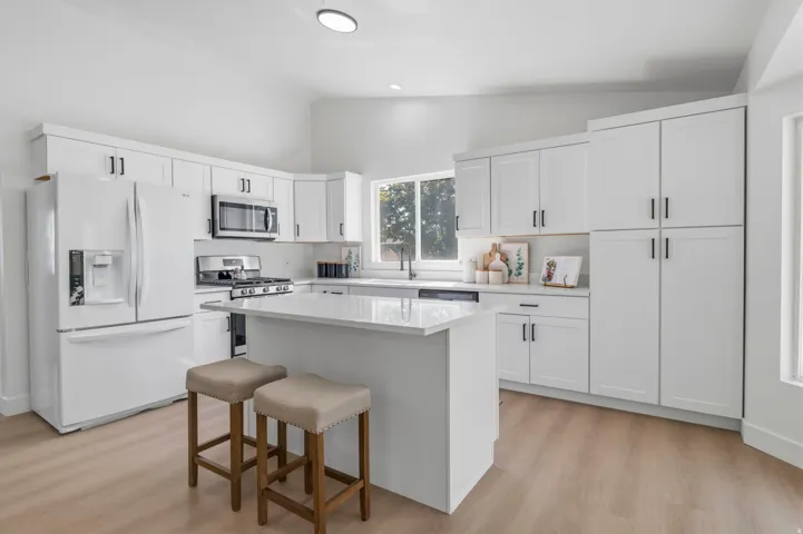 Modern kitchen featuring white shaker cabinetry, stainless steel appliances, a center island with a countertop, wood-finish flooring, and a large window