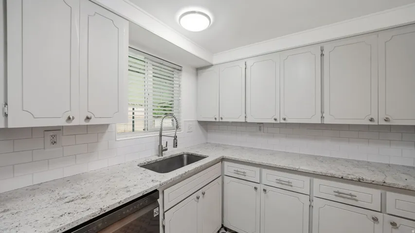Bright kitchen featuring white cabinetry with decorative trim, light granite-style countertops, white subway tile backsplash, a stainless steel sink, and a modern pull-down faucet
