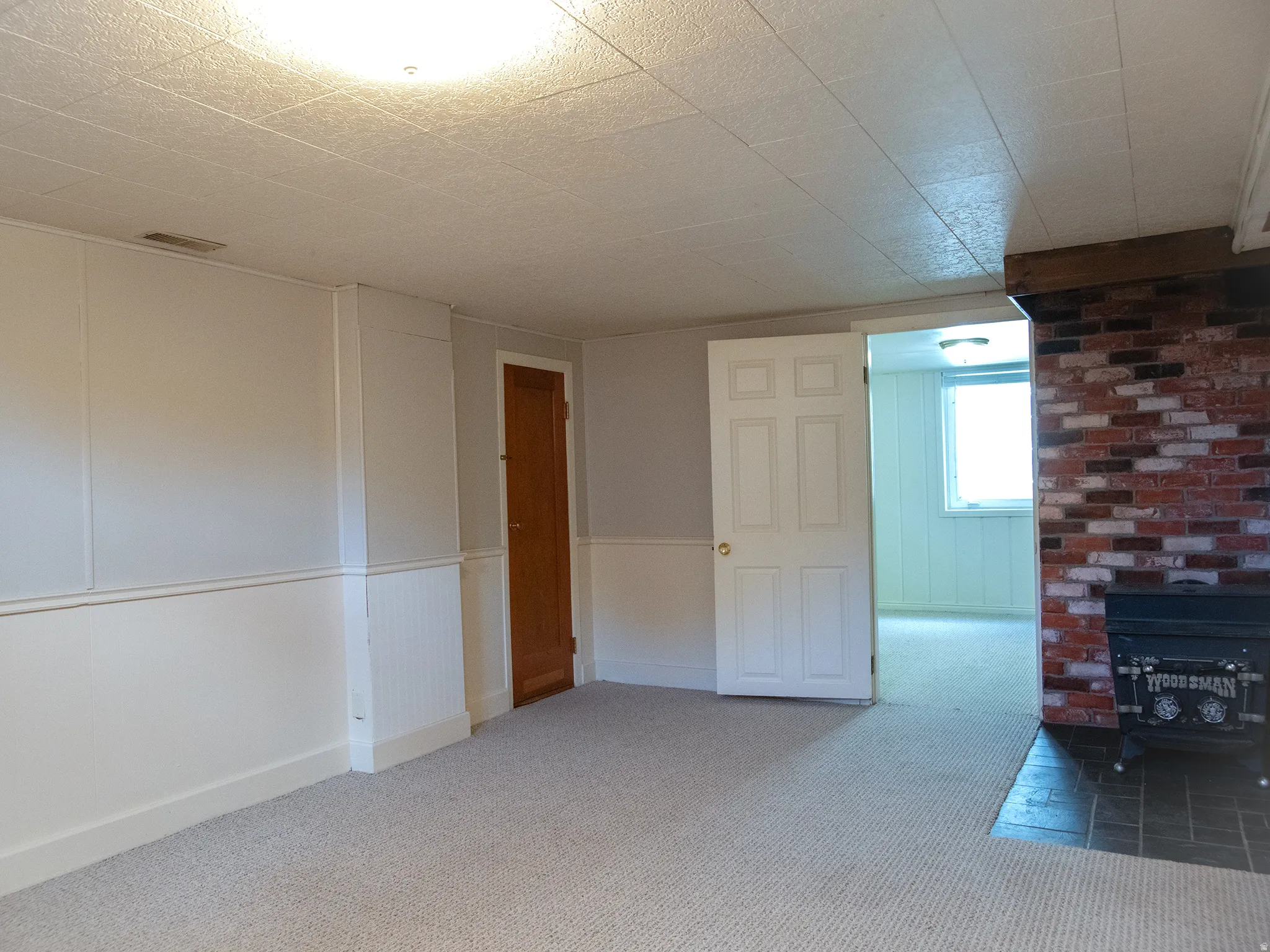 Brick accent wall with a black wood stove, carpeted flooring, and a white paneled ceiling