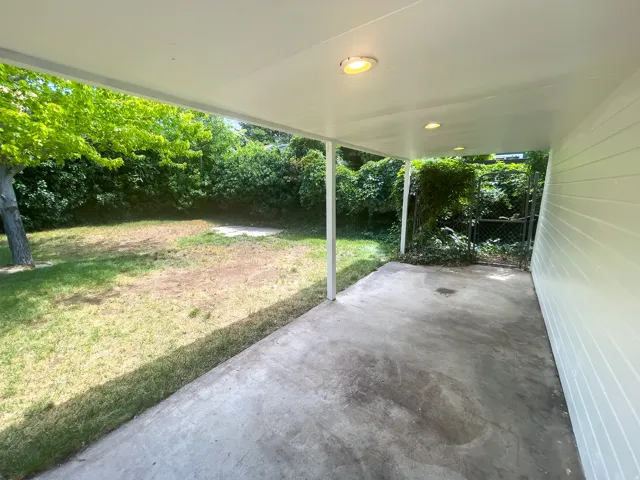 Covered patio with integrated ceiling lighting, concrete surface, and direct access to a grassy yard featuring mature trees and a chain-link gate