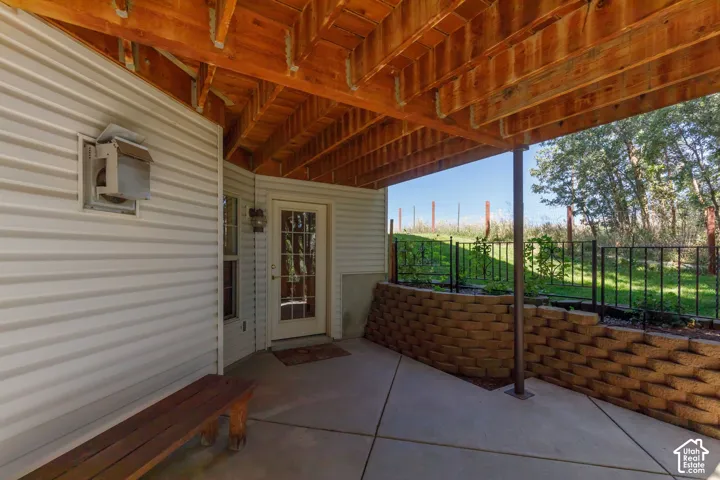 Concrete patio with exposed wood ceiling, white siding, a glass-panel door, and a retaining wall with a black metal fence