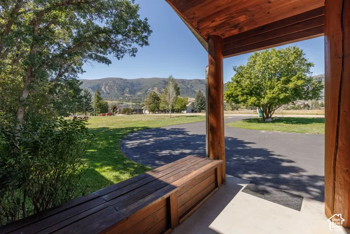 Covered porch with wood-finish columns and ceiling, built-in wooden bench, concrete flooring, and an expansive lawn with mature trees and a paved driveway