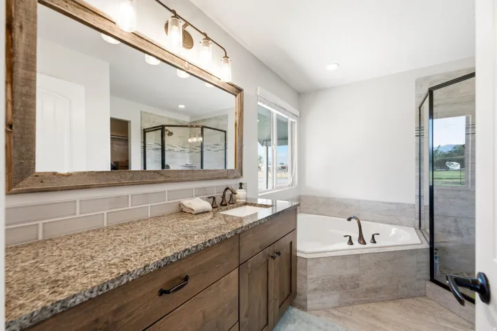 Granite countertop with an undermount sink, wood-finish vanity, a large framed mirror, a corner soaking tub, and a glass-enclosed shower