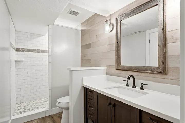Walk-in shower with subway tile and pebble floor, dark wood-finish vanity with countertop, framed mirror, and wood-finish accent wall