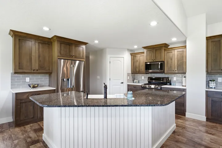 Kitchen featuring wood-finish cabinetry, stainless steel appliances, a center island with a built-in sink, stone countertops, and a subway tile backsplash