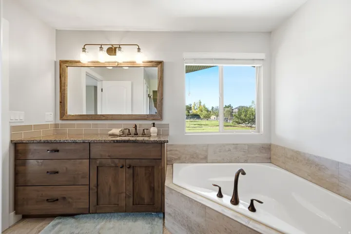 Built-in corner bathtub with dark bronze fixtures, wood-finish vanity with granite countertop, undermount sink, and a framed mirror with overhead lighting