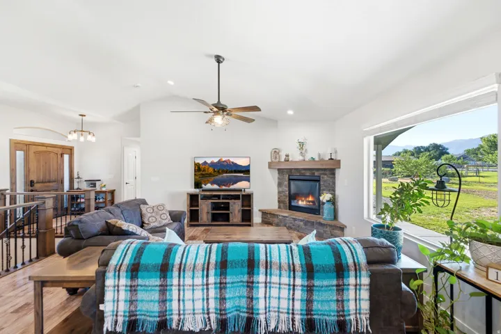 Vaulted ceiling, wood-finish flooring, stone-clad fireplace with wood mantel, large picture window, and ceiling fan with integrated lighting