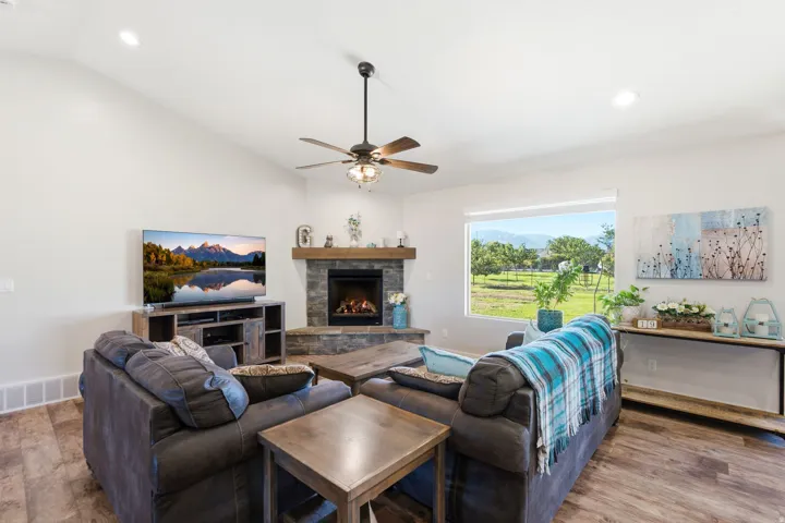 Vaulted ceilings with recessed lighting, wood-finish flooring, stone-clad fireplace with wood mantel, large window with outdoor scenery, and a ceiling fan