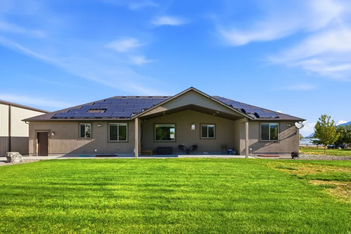 Expansive grassy yard with a covered patio, solar panels, stucco exterior, and a visible body of water in the background
