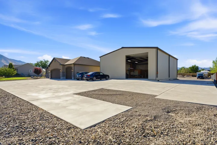 Expansive concrete driveway, detached metal outbuilding with large bay door, attached two-car garage, stucco exterior with stone accents, and mature landscaping