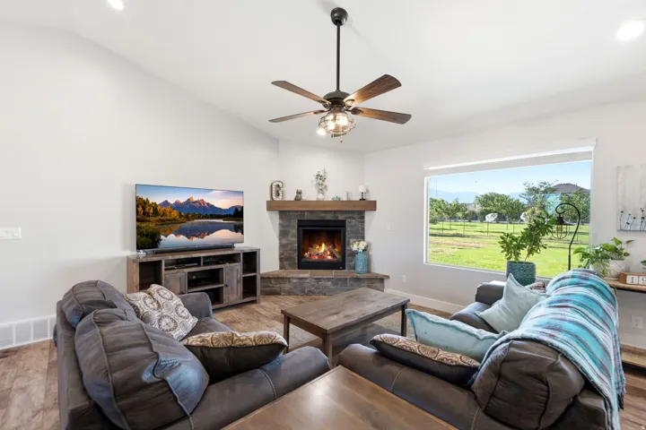 Vaulted ceiling with a ceiling fan, corner fireplace with stone surround and wood mantel, large window, and wood-finish flooring