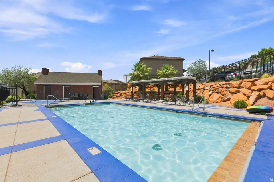 Resort-style pool area featuring a rectangular pool with blue tile accents, a covered pergola with lounge seating, and a red rock landscape wall