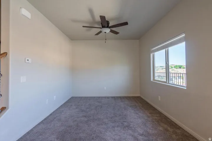 Carpeted interior space featuring a ceiling fan with integrated lighting, a large window with roller shades, and light-toned wall paint