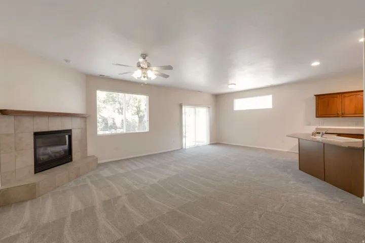 Spacious living area featuring a tiled fireplace with wood mantel, large window with blinds, ceiling fan with light fixture, and sliding glass door