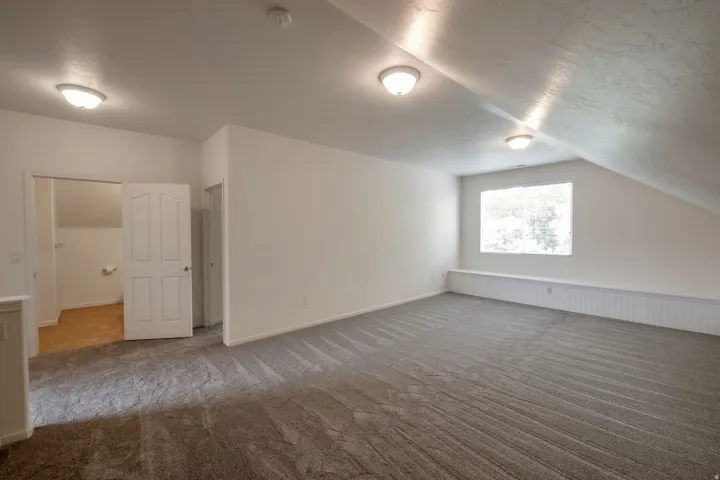 Spacious attic room featuring sloped ceilings, plush gray carpet, a white window seat with paneling, and three flush-mount dome light fixtures