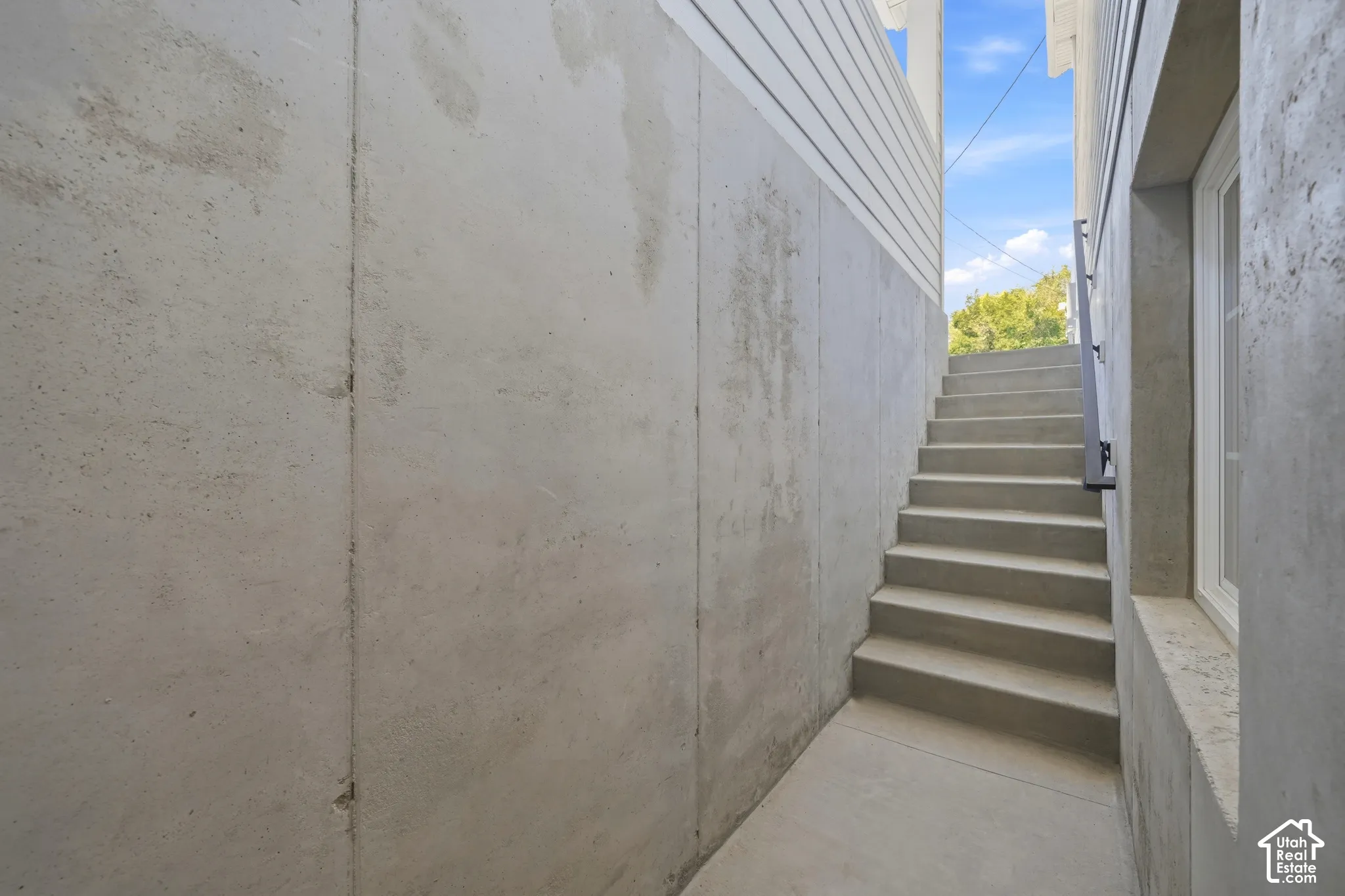 Concrete exterior staircase with a dark metal handrail, bordered by concrete walls and a building with white horizontal siding