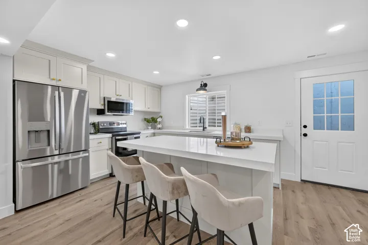 Modern kitchen featuring a large central island, stainless steel appliances, recessed lighting, wood-finish flooring, and crisp white cabinetry with gold-tone hardware
