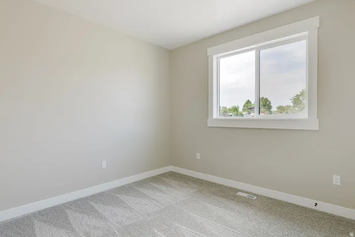 Well-lit room featuring a large window with white trim, light neutral wall paint, and patterned gray carpeting