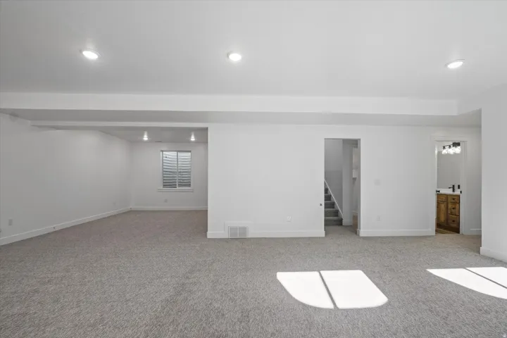 Spacious lower-level room featuring light gray carpeting, recessed lighting, white walls, and a window with horizontal blinds