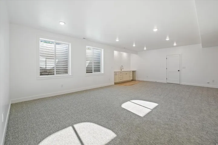 Spacious lower-level room featuring light gray carpeting, two egress windows, a built-in wet bar with light cabinetry and a stainless steel sink, wood-finish flooring at the bar, and recessed lighting