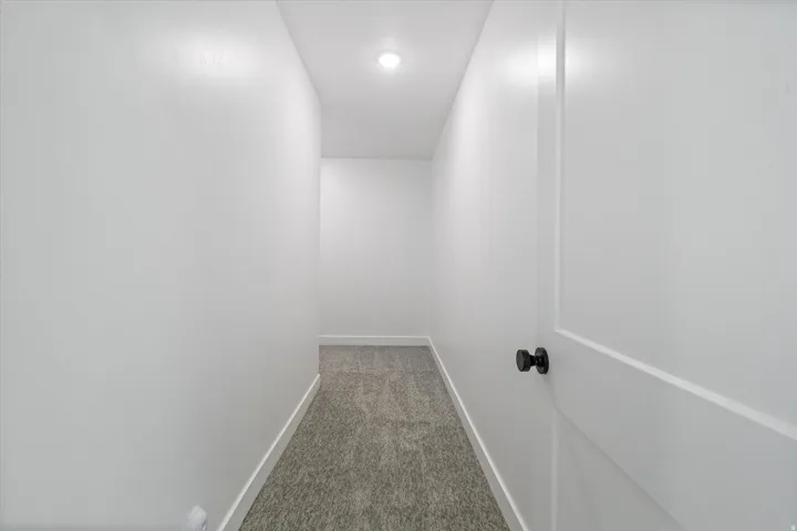 Recessed ceiling lighting illuminates a hallway featuring white walls, white baseboards, and patterned gray carpeting