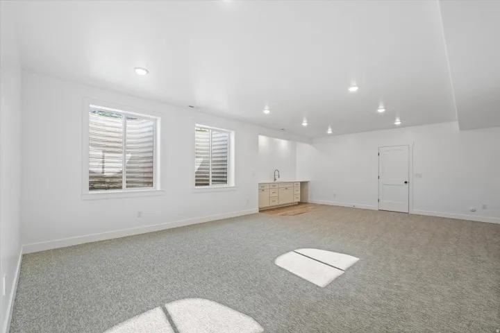 Spacious room featuring light gray carpet flooring, two windows, recessed lighting, and a built-in wet bar with a sink and light-toned cabinetry