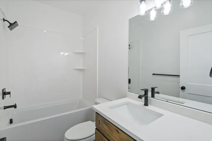 Bathroom featuring a white tub/shower combination with integrated shelving, a white toilet, a wood-finish vanity with a white countertop and integrated sink, and a large mirror with a multi-bulb vanity light fixture