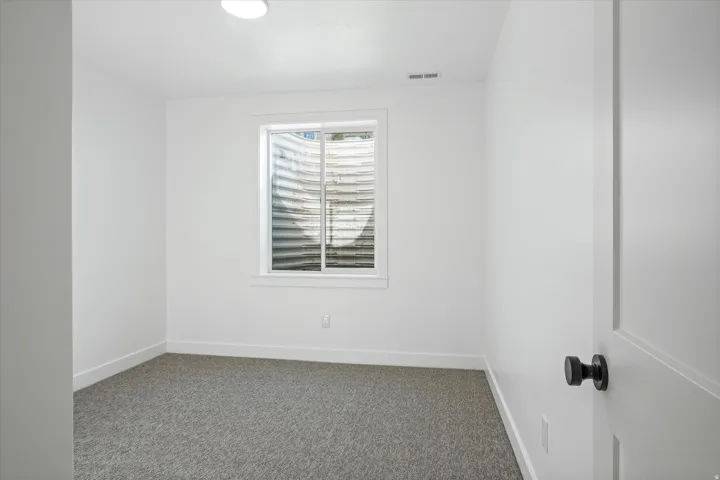 Bright room featuring a window with a corrugated metal window well, light gray carpet, white walls, and a white paneled door with a matte black knob