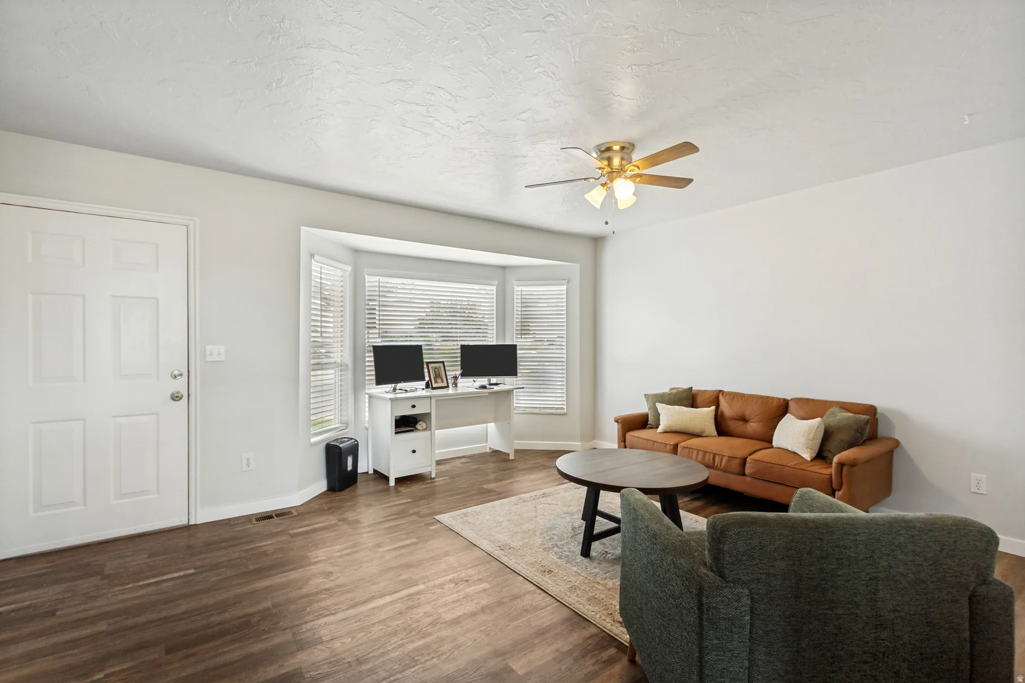 Living area with wood-finish flooring, white walls, and a multi-pane bay window with blinds