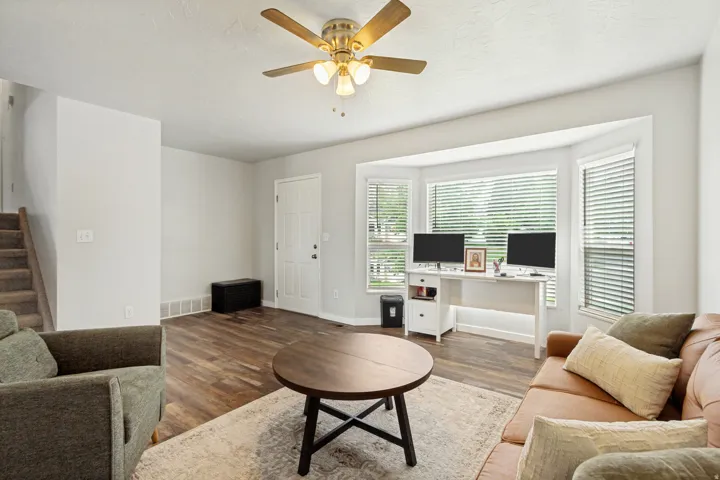 Living area featuring wood-finish flooring, a ceiling fan with integrated lighting, and a bay window with horizontal blinds